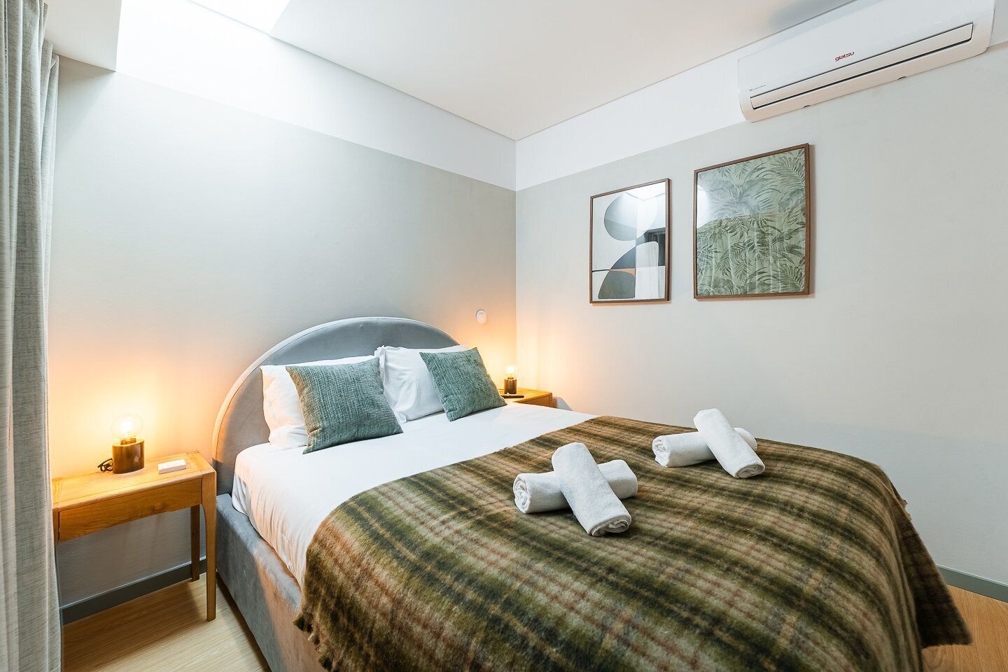 A hotel room with a bed, bedside tables, an air conditioner, and two framed artworks on the wall.