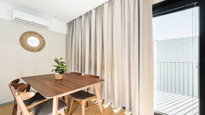 Une pièce comprenant une table à manger en bois, quatre chaises, une plante en pot, un miroir rond et une porte-fenêtre coulissante donnant sur un balcon.