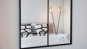A modern bedroom with a bed, pillows, a nightstand, and a mirror reflecting a lamp and a towel. - GuestReady - Modern ambience in Porto (Porto)