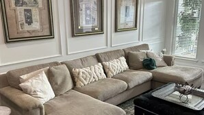 A beige sectional sofa with multiple cushions, a black ottoman, and a glass coffee table with decorative items. - Lovely 4-bedroom house in charming Irving with AC, WiFi - Coppell Schools. (Irving)
