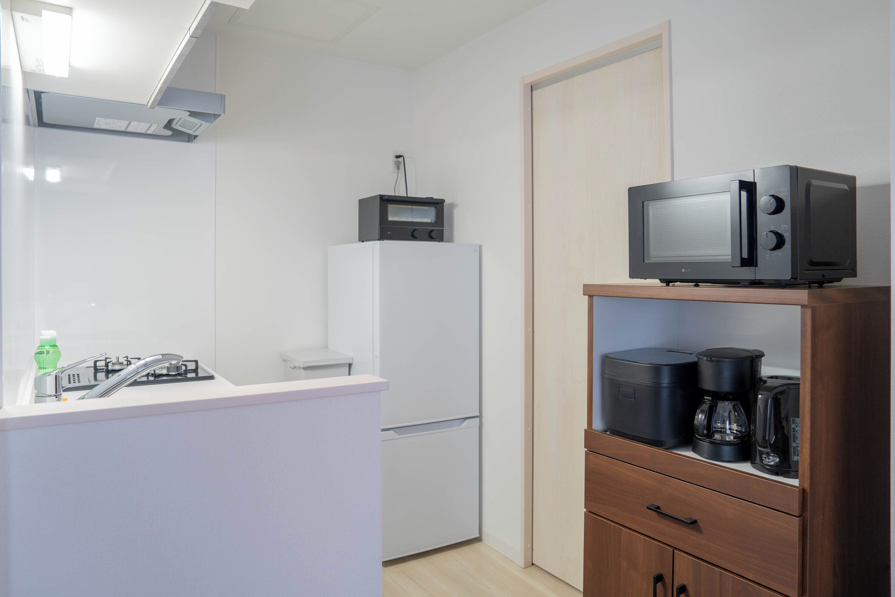 A compact kitchen with a white countertop, a built-in microwave, a small refrigerator, and a coffee maker on a wooden shelf.