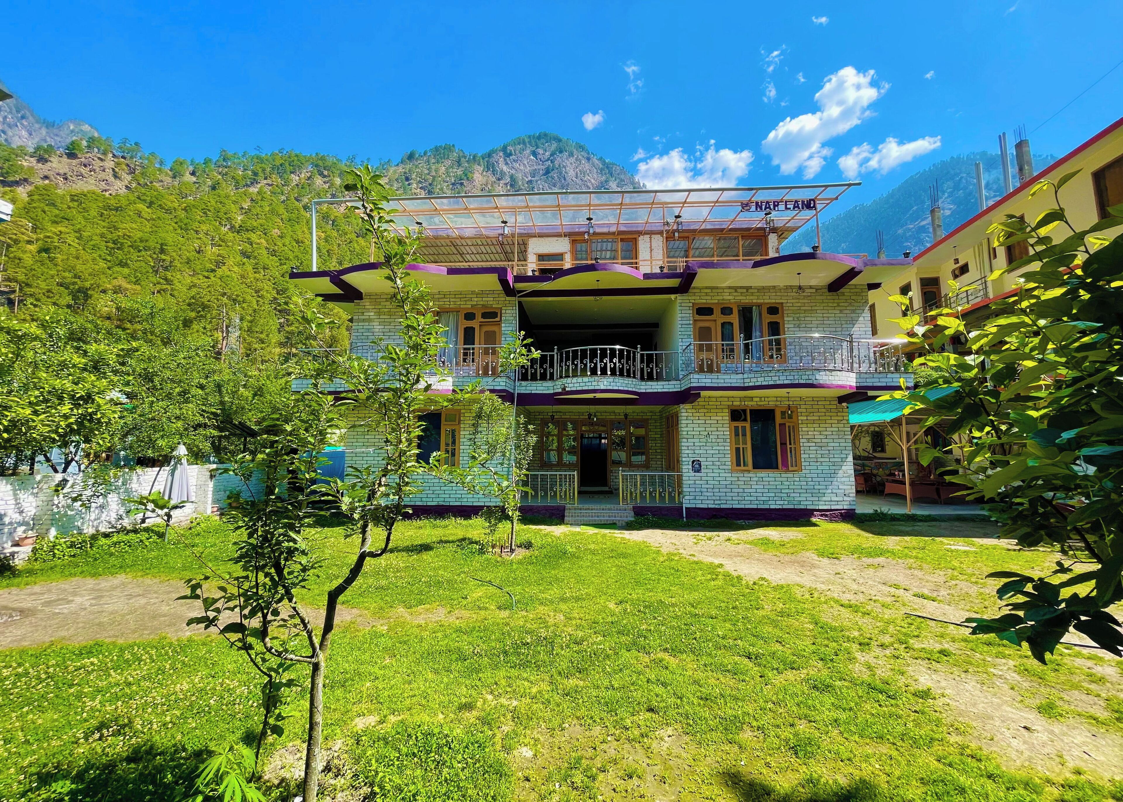 A two-story building with a balcony, surrounded by greenery and mountains in the background.