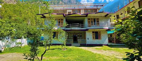 A two-story building with a balcony, surrounded by greenery and mountains in the background.