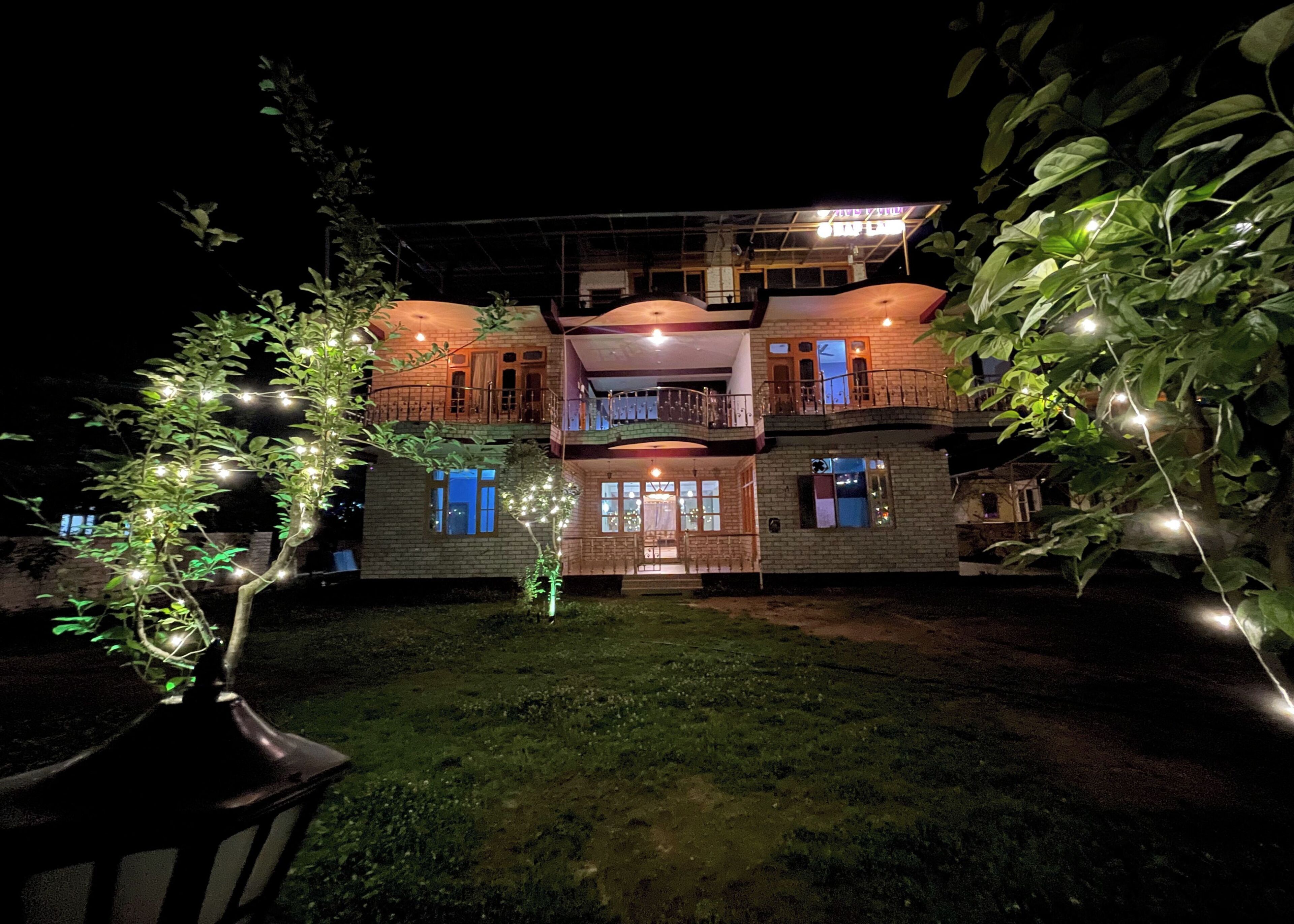 A two-story building with a balcony on the upper floor, surrounded by greenery and string lights.