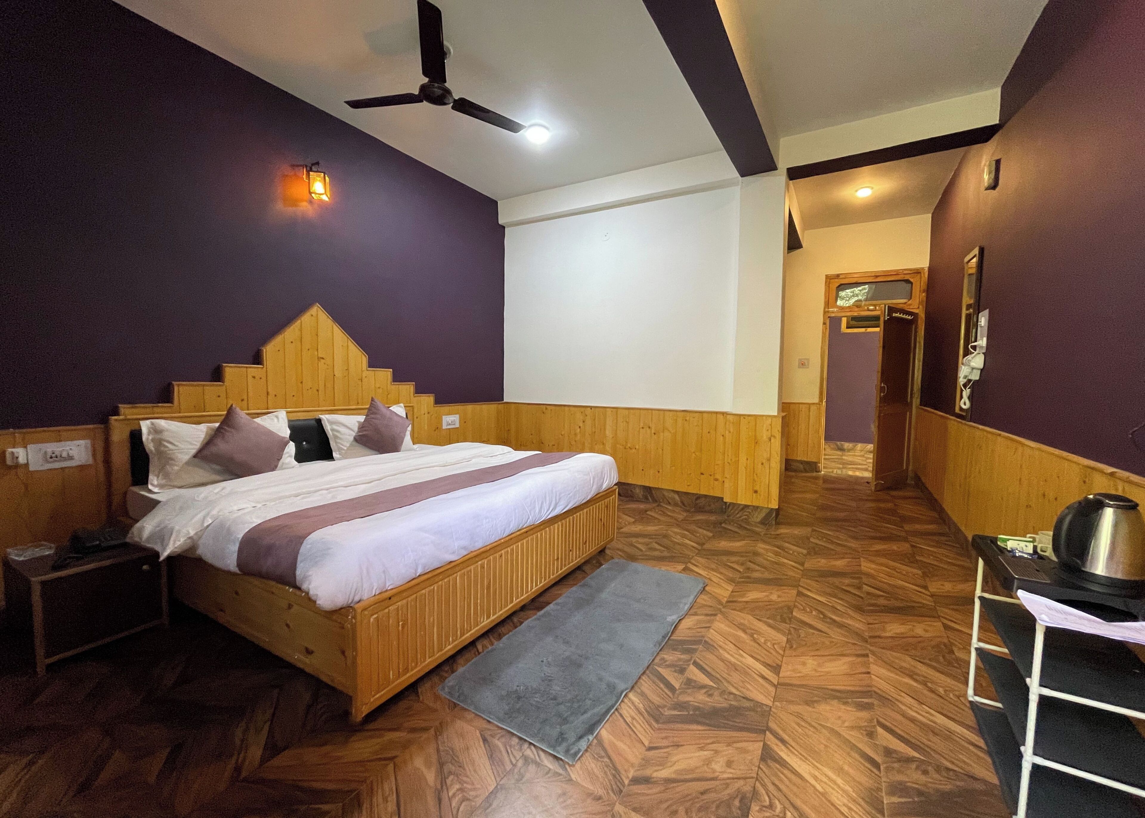 A bedroom with a wooden bed, a nightstand, a desk with a kettle, and a ceiling fan.