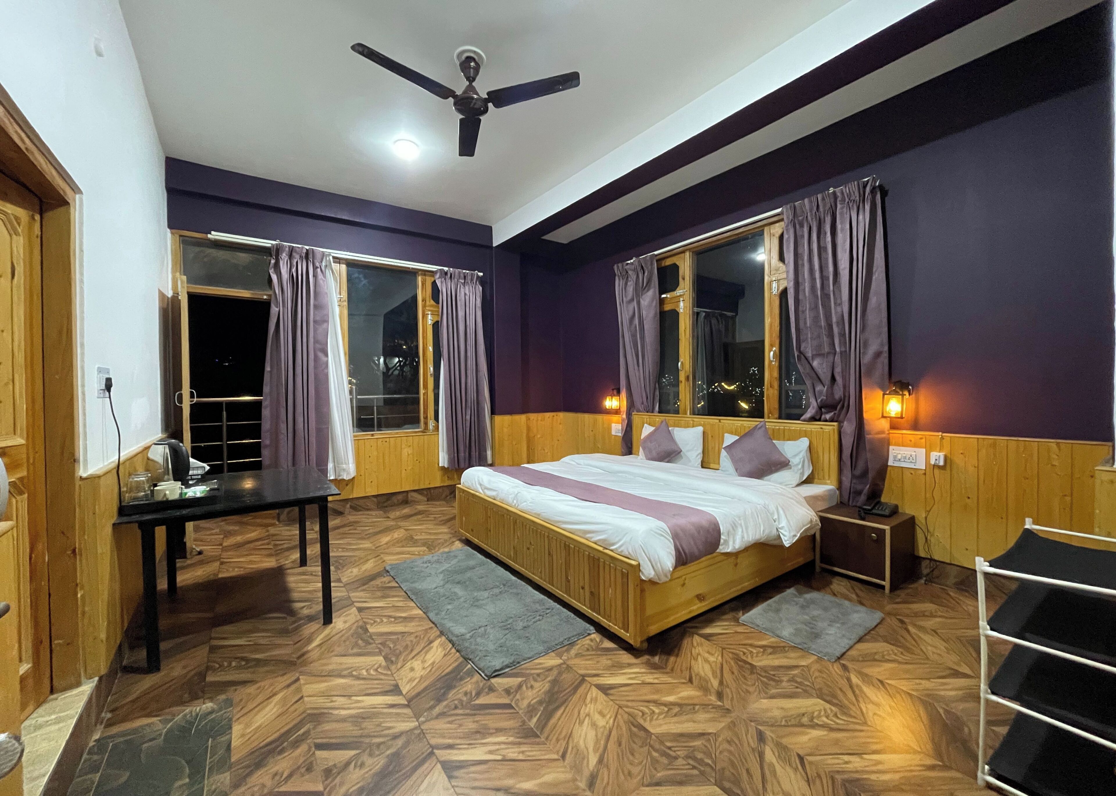 A bedroom with a bed, wooden floor, ceiling fan, window with curtains, bedside table, and nightstand.