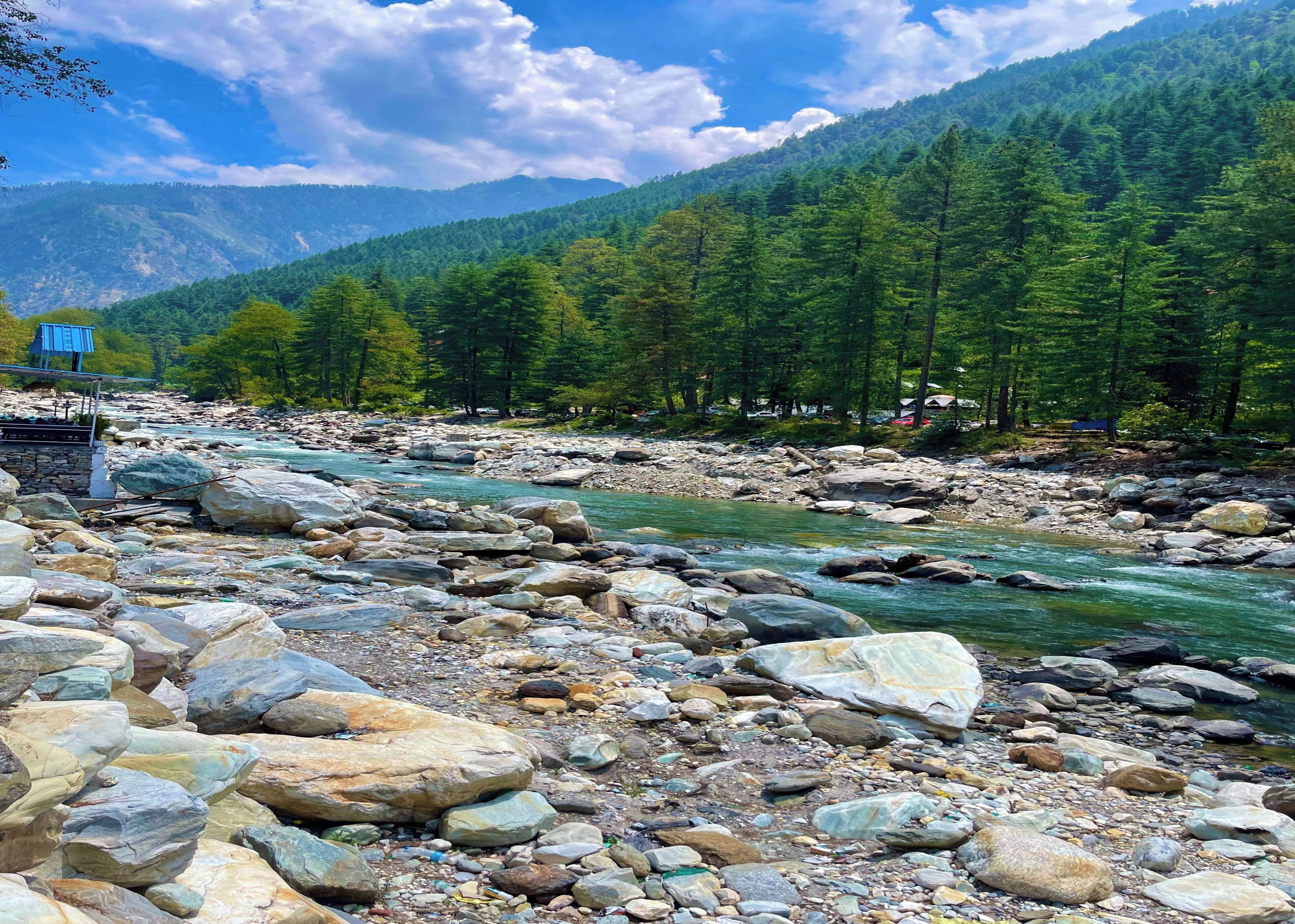 A river with large rocks, a wooden bridge, and a forested mountain in the background.