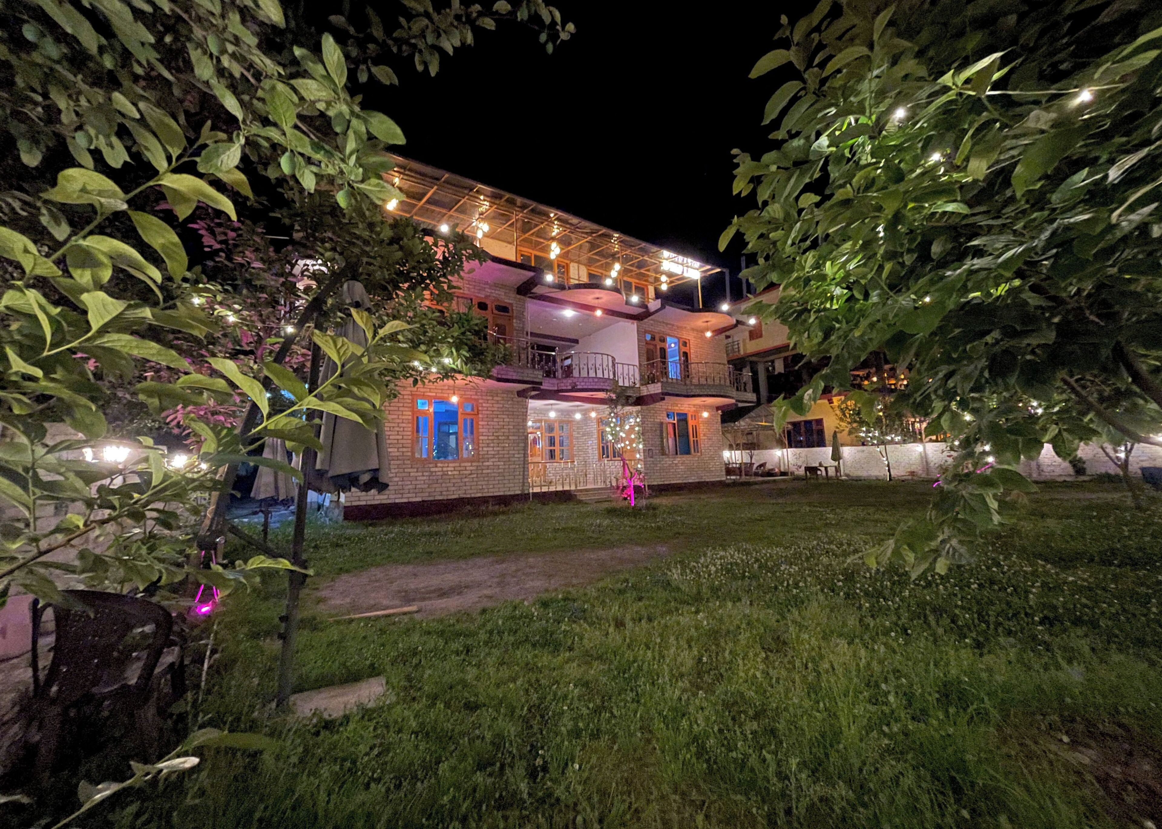 A multi-story building with a balcony, surrounded by greenery and illuminated by string lights.