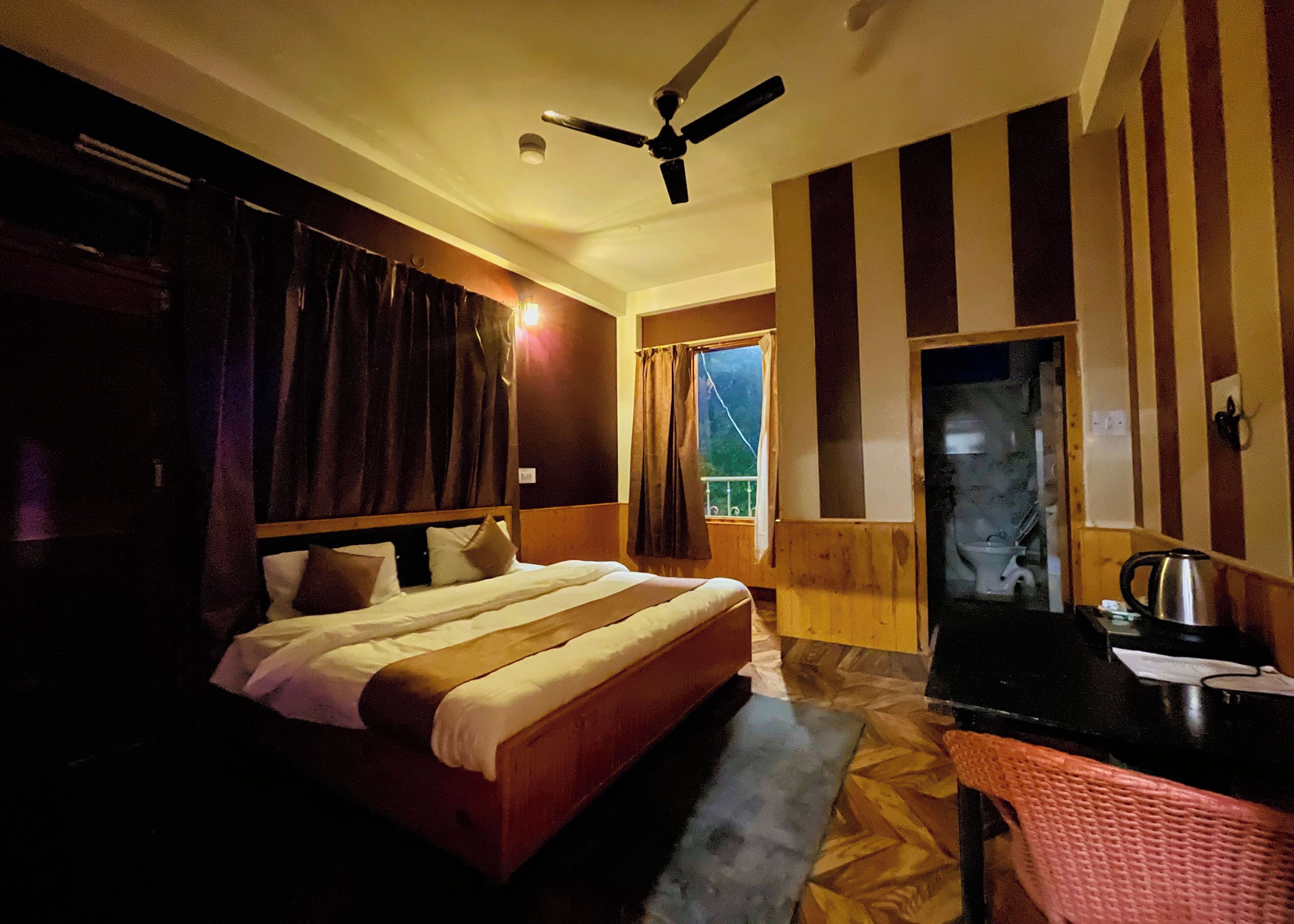 Une chambre d’hôtel avec un lit, un bureau doté d’une bouilloire, un ventilateur de plafond et une fenêtre avec des rideaux.