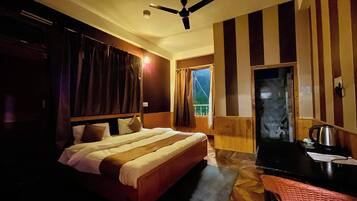 A hotel room with a bed, a desk with a kettle, a ceiling fan, and a window with curtains.