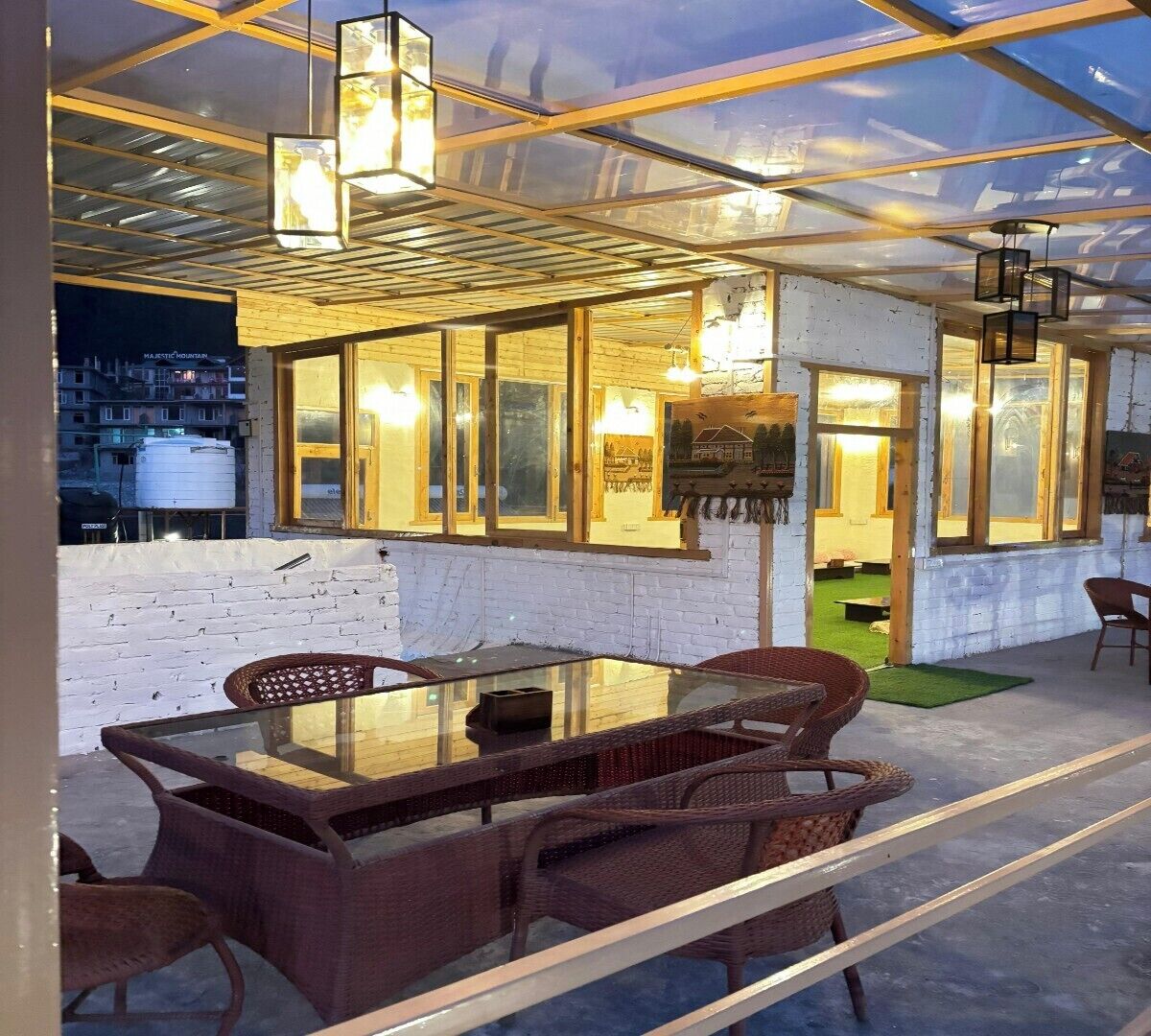 Un espacio de comedor al aire libre con mesa de vidrio y sillas de mimbre, rodeado por una estructura de cristal con vigas de madera y luces colgantes.