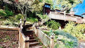 Un escalier en bois mène à une terrasse en bois, bordée d’un mur de pierres et agrémentée de verdure.