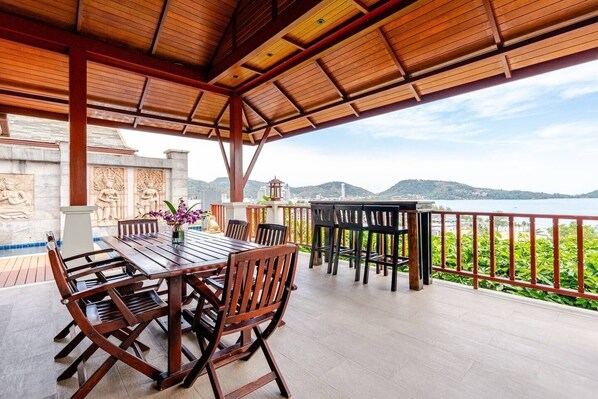 Un espacio de comedor al aire libre cubierto, con mobiliario de madera y vistas a una masa de agua y colinas.