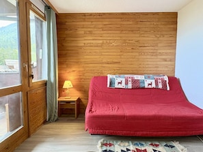 A room with a wooden wall, a bed with a red blanket, a small table with a lamp, and a view through a glass door.