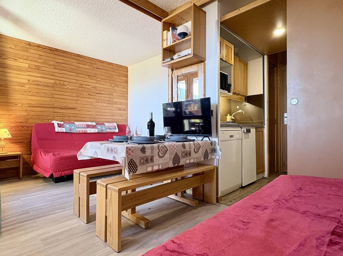 A compact living space with a red sofa, a wooden bench, a dining table with a patterned tablecloth, a television, and a kitchenette.