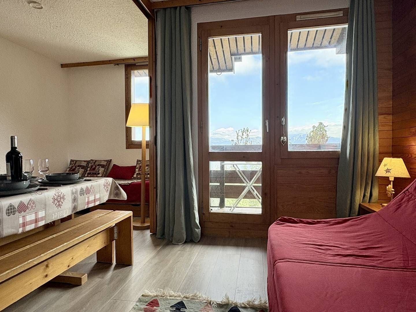 Una habitación acogedora con un banco de madera, una mesa lista para comer, un sofá rojo y vistas a través de una puerta de madera.