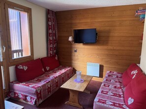 A cozy room with a wooden wall, a television mounted above a table, and two sofas with heart-patterned covers.