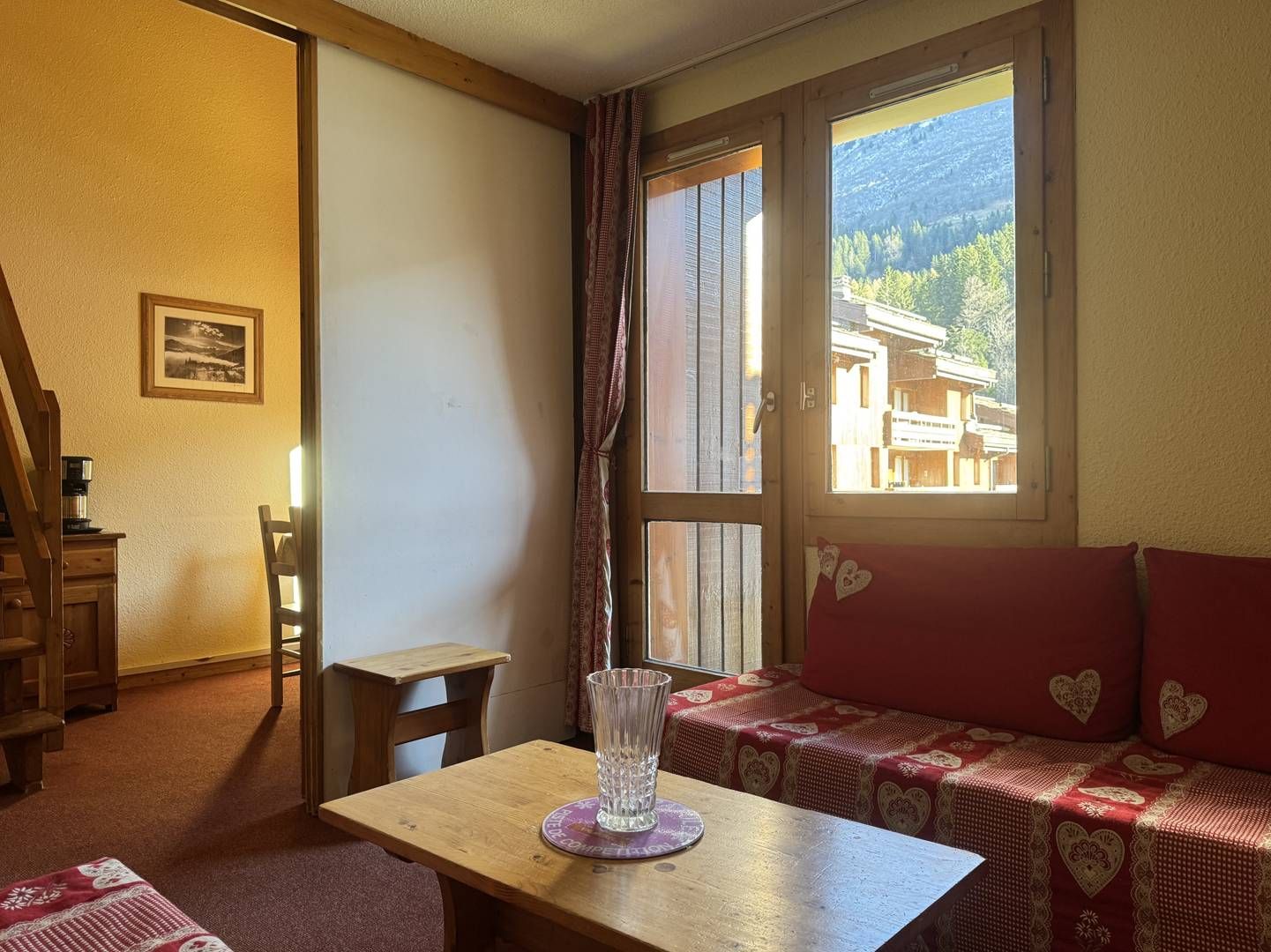 Une pièce chaleureuse avec une table en bois, un canapé rouge orné de motifs en forme de cœur, une fenêtre donnant sur des bâtiments et des montagnes, et un escalier.