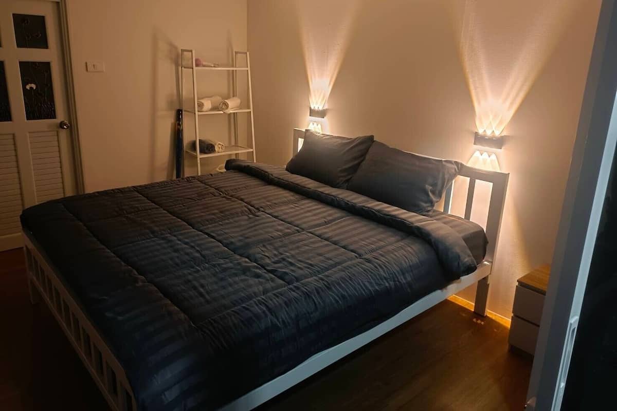 A neatly made bed with a dark quilt, a white headboard, and two wall-mounted lamps.