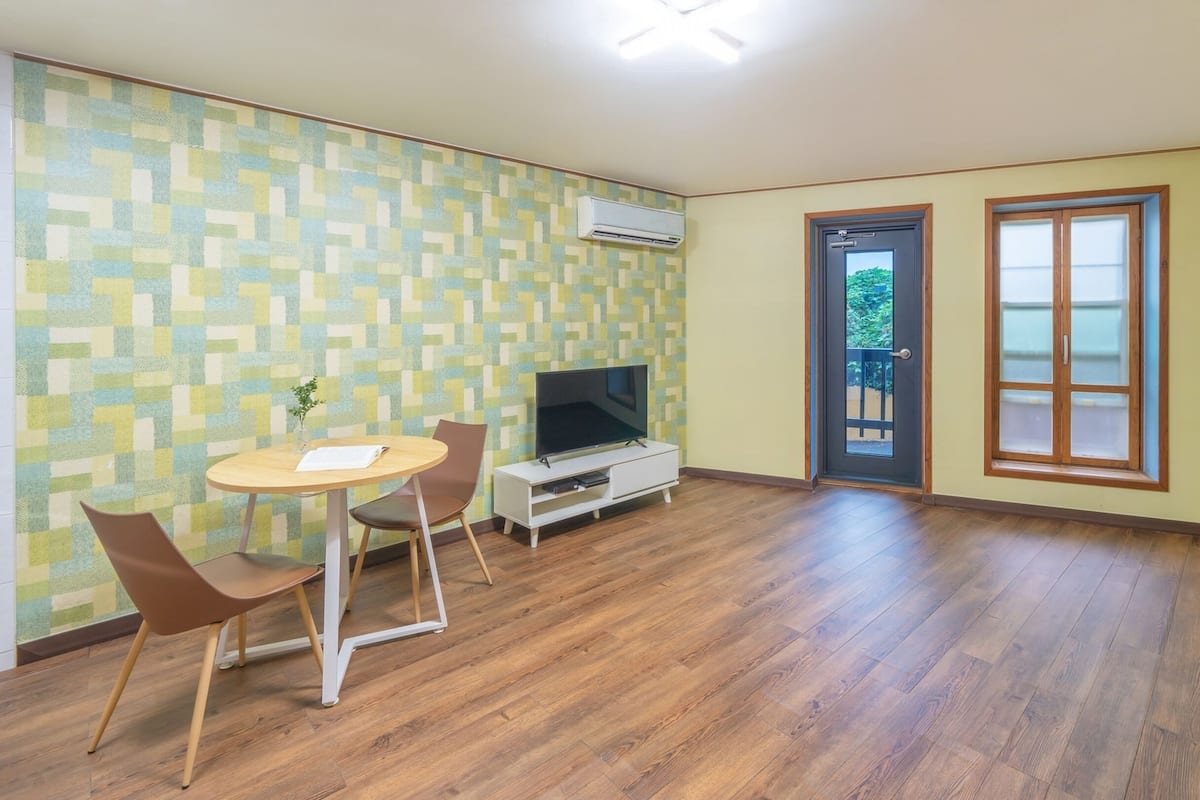 Une pièce avec un plancher en bois, un téléviseur posé sur un meuble blanc, deux chaises, une petite table et un mur orné d’un motif.
