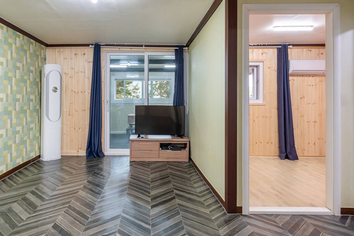 Une pièce dotée d’un parquet à motifs en chevrons, d’un téléviseur posé sur un meuble en bois et d’une porte-fenêtre coulissante donnant sur un espace extérieur.