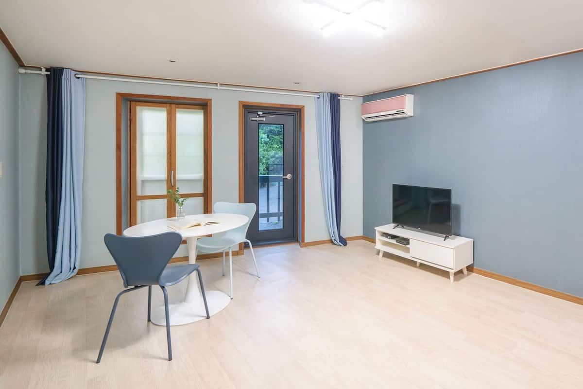 Une pièce comprenant une table à manger, deux chaises, un téléviseur posé sur un meuble blanc et une porte donnant sur l’extérieur.
