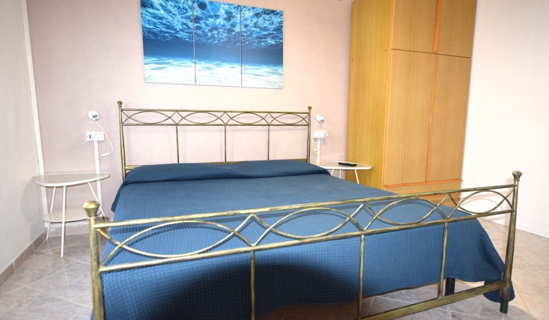 Uma cama com estrutura metálica e roupa de cama azul, uma pequena mesa de cabeceira, uma luminária fixada na parede e um guarda-roupa de madeira.