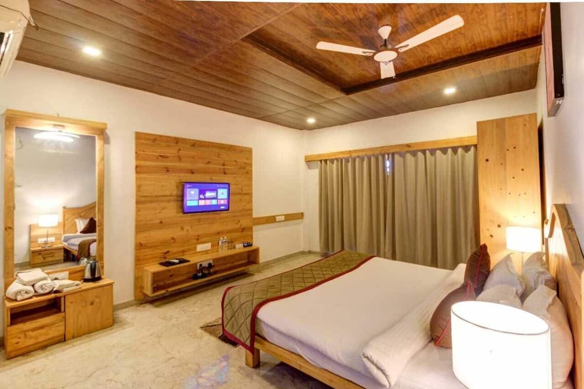 A hotel room with a wooden ceiling, a bed with a quilted bedspread, a television mounted on the wall, a wooden bench, a bedside table with a lamp, and a window with curtains.