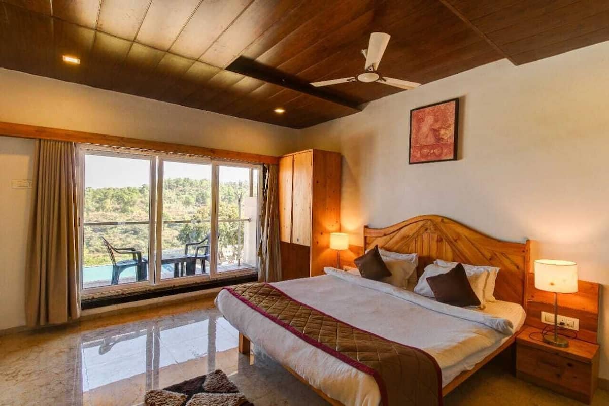 A bedroom with a large bed, wooden headboard, two bedside lamps, a ceiling fan, and a view through a sliding glass door to an outdoor area with chairs.
