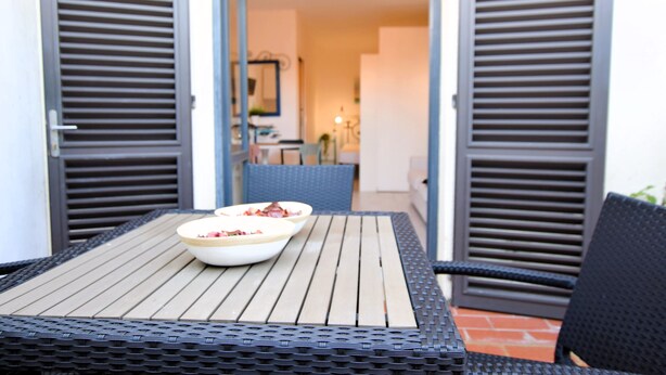 Un espacio de comedor al aire libre con una mesa y sillas, un tazón de comida y vistas del interior a través de contraventanas abiertas.