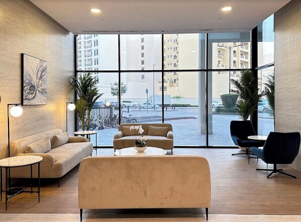A modern lounge area with a sofa, armchairs, and a coffee table. Large windows offer a view of the street and buildings.