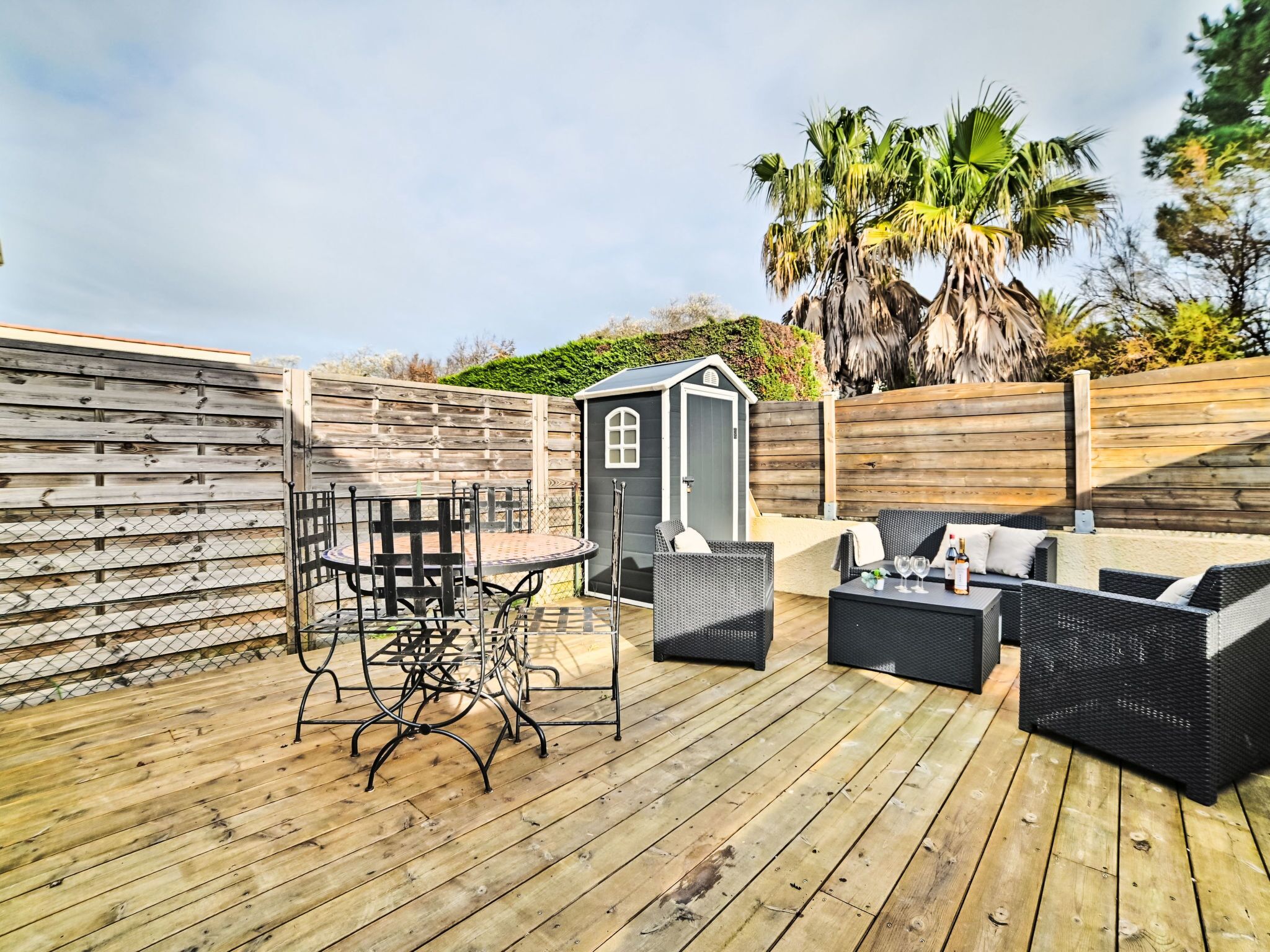 Une terrasse en bois avec des meubles d’extérieur, un petit abri de jardin et un palmier.