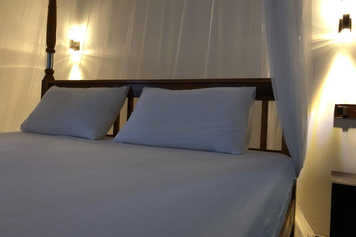 A bed with white linens and a wooden headboard, a nightstand with a lamp, and a wall-mounted light fixture.