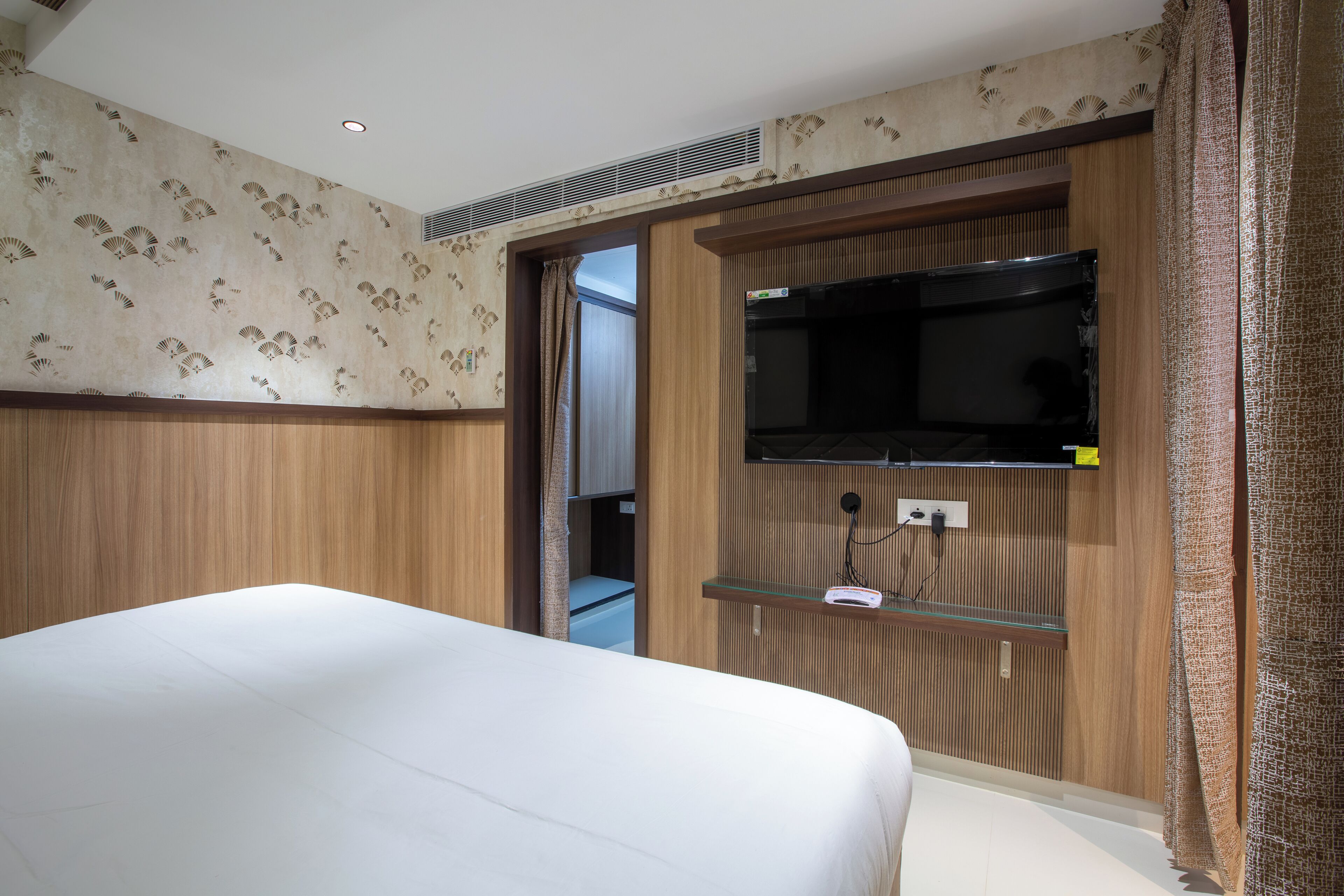 A hotel room with a bed, a TV mounted on a wooden panel, and a small shelf.