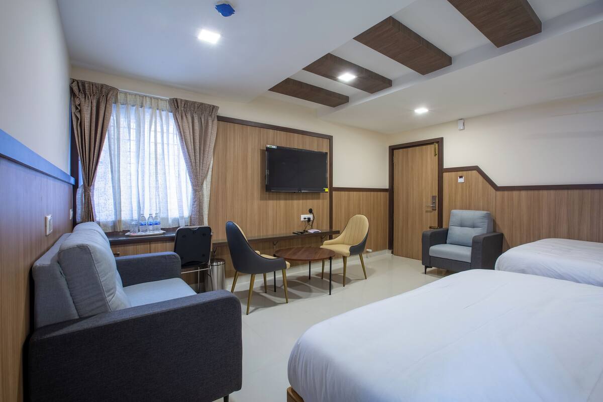 Habitación de hotel con sofá, dos camas individuales, escritorio con silla, televisión montada en la pared y ventana con cortinas.