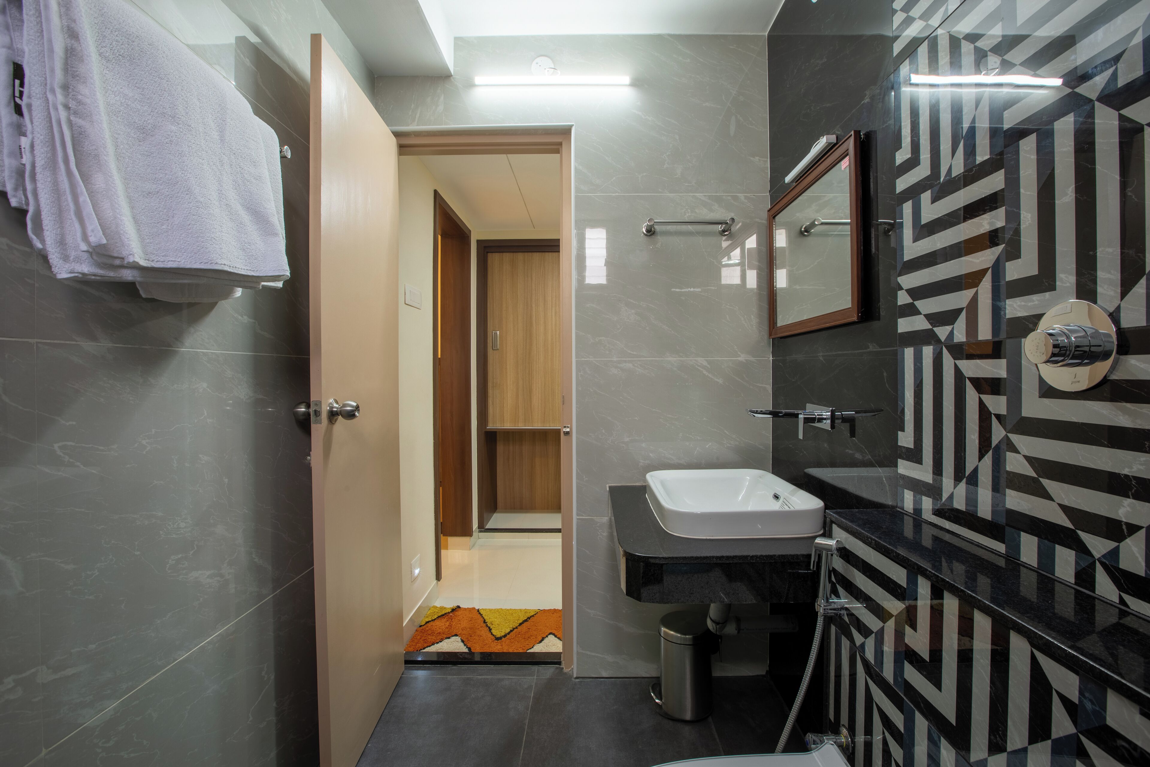 A modern bathroom with a vanity, sink, and mirror. There is a towel rack with towels, a trash can, and a patterned floor.