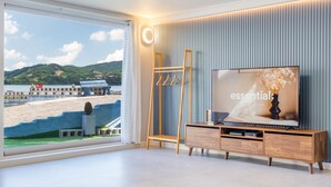 A modern living room with a large window overlooking a beach, a wooden TV stand, and a wall-mounted TV displaying 'essential'. - Namhae Yujane Pension (Namhae)