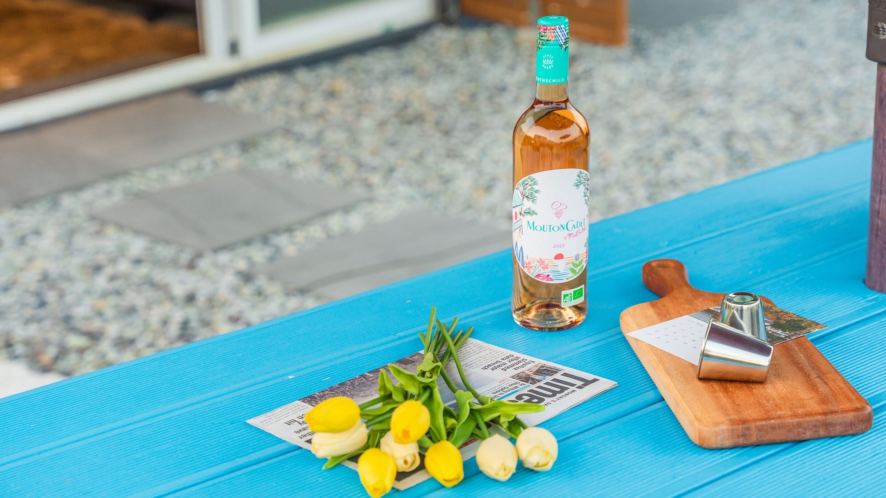Una botella de vino Mouton Cadet 2022, una tabla de madera con un cuchillo y un ramo de flores amarillas y blancas sobre una mesa azul.