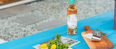 Una botella de vino Mouton Cadet 2022, una tabla de madera con un cuchillo y un ramo de flores amarillas y blancas sobre una mesa azul.