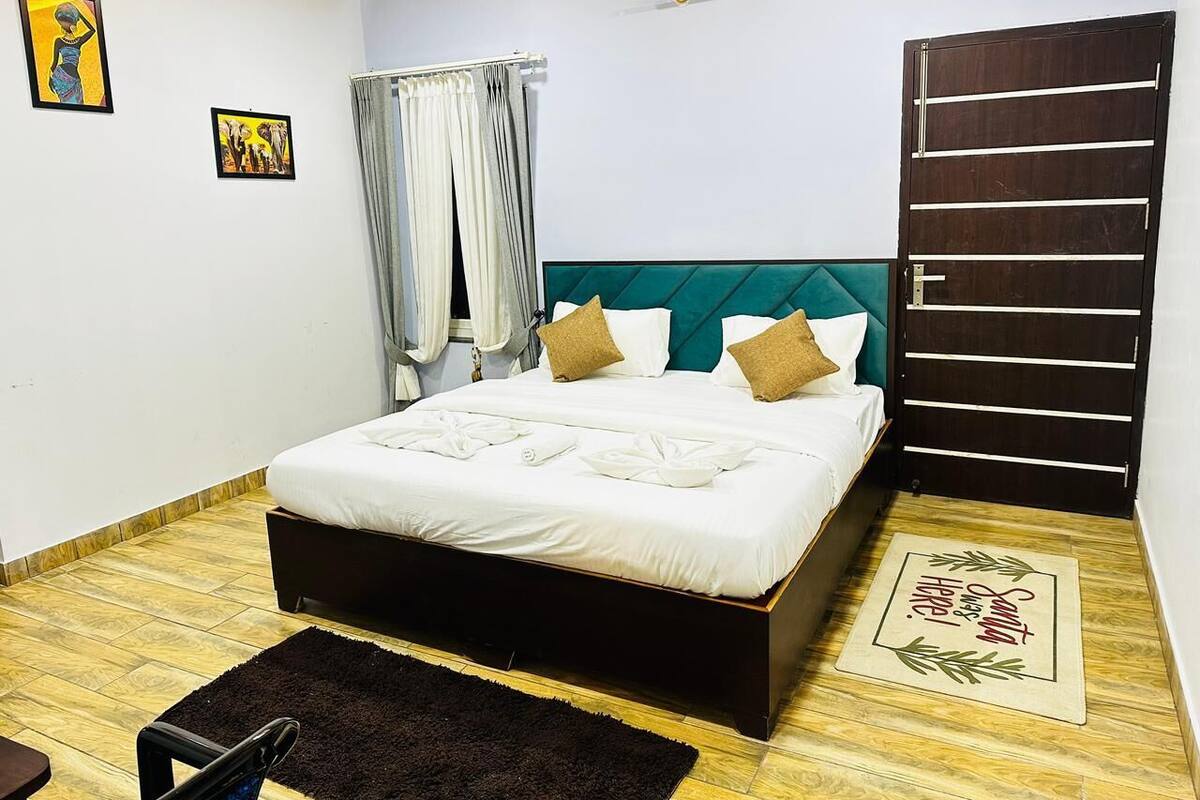 Ein Hotelzimmer mit Bett, schwarzem Teppich, Holzboden, weißer Wand mit zwei gerahmten Bildern, Fenster mit Vorhängen und einer Tür mit horizontalen Linien.