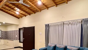 Un salon spacieux avec un canapé d’angle, des poufs et une table basse en verre. La pièce est dotée d’un plafond en bois avec des poutres apparentes et d’un ventilateur de plafond.