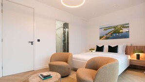 A modern hotel room with a large bed, two armchairs, a small table, a glass shower enclosure, and a painting of a coastal landscape. - LevelUp Sunrise (Amsterdam)