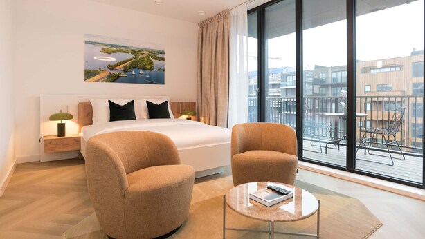 A modern hotel room with a large bed, two armchairs, a small round table, and a large window offering a view of the city. - LevelUp Sunrise (Amsterdam)