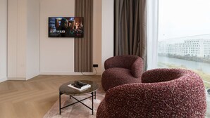 Un salon moderne avec un téléviseur fixé au mur, une table basse ronde sur laquelle repose un livre, un fauteuil rouge recouvert de tissu, et une vue sur le paysage urbain à travers de grandes fenêtres.