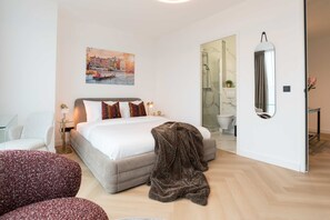 A modern hotel room with a bed, a chair, a round ottoman, a painting of a cityscape, a shower area, and a mirror.