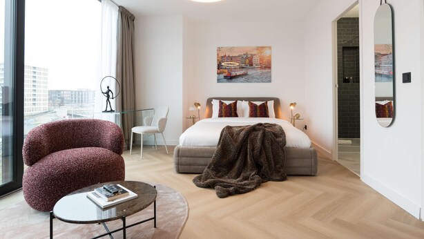 Une chambre d’hôtel moderne avec un grand lit, un coin salon comprenant un fauteuil et une table, une vue sur la ville par la fenêtre et un tableau représentant un pont accroché au mur.
