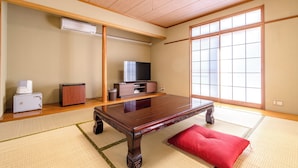 Ein traditioneller japanischer Raum mit Tatami-Bodenbelag, einem Holztisch, einem Fernseher und einer Schiebetür.