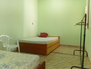Una habitación pequeña con cama individual, cama de madera, una silla de plástico blanco y un atril metálico negro.