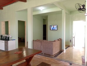 Una habitación con una mesa de madera, un televisor montado en la pared y una zona de estar con cojines.