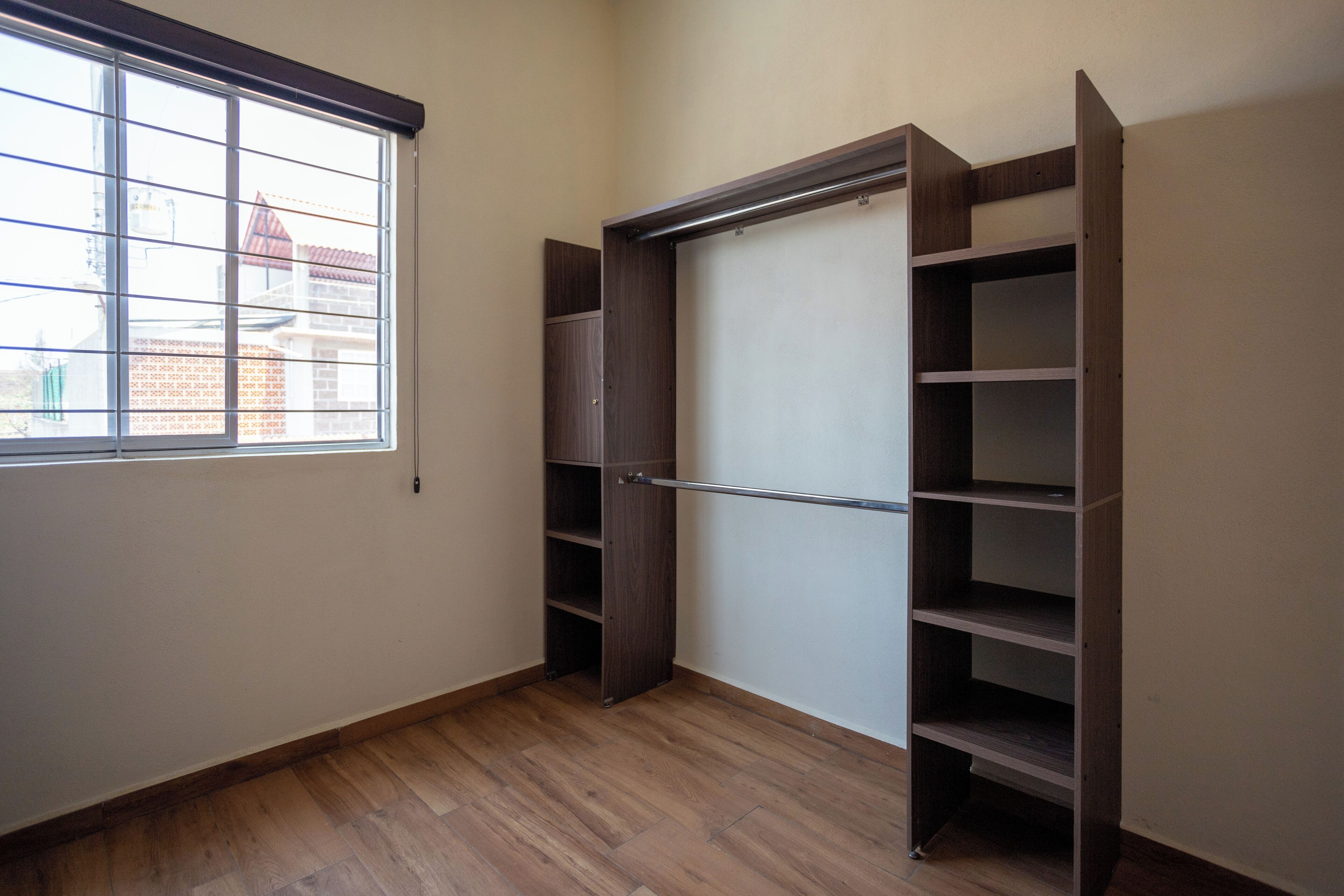 Una habitación con piso de madera, ventana con persianas y armario empotrado con varios estantes.